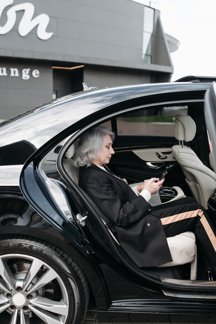 Senior woman in business attire sits in a luxury car, using a tablet outside a modern building.
