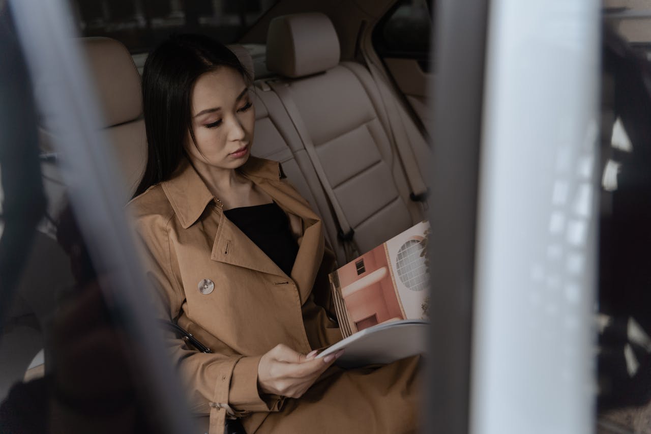 Well-dressed Asian woman reading a magazine in a car interior captured from the side view.